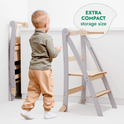 A small child stands near the Toddler Tower: Foldable Step Stool for Toddlers - Gray in a white room. One stool is open while another is folded neatly against the wall. Text reads: EXTRA COMPACT storage size, making this foldable step stool ideal for saving space when not in use.