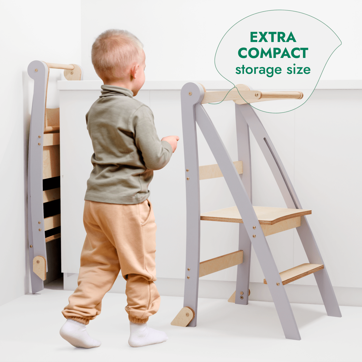 A small child stands near the Toddler Tower: Foldable Step Stool for Toddlers - Gray in a white room. One stool is open while another is folded neatly against the wall. Text reads: EXTRA COMPACT storage size, making this foldable step stool ideal for saving space when not in use.