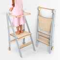 A child in pink stands on a Toddler Tower: Foldable Step Stool for Toddlers - Gray, featuring safety rails and a light blue and wood finish. Another identical foldable step stool is folded beside it, showcasing its compact, space-saving design.