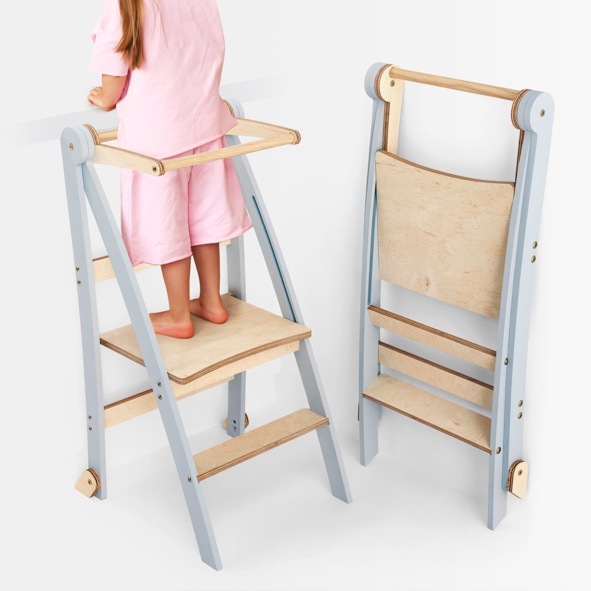 A child in pink stands on a Toddler Tower: Foldable Step Stool for Toddlers - Gray, featuring safety rails and a light blue and wood finish. Another identical foldable step stool is folded beside it, showcasing its compact, space-saving design.