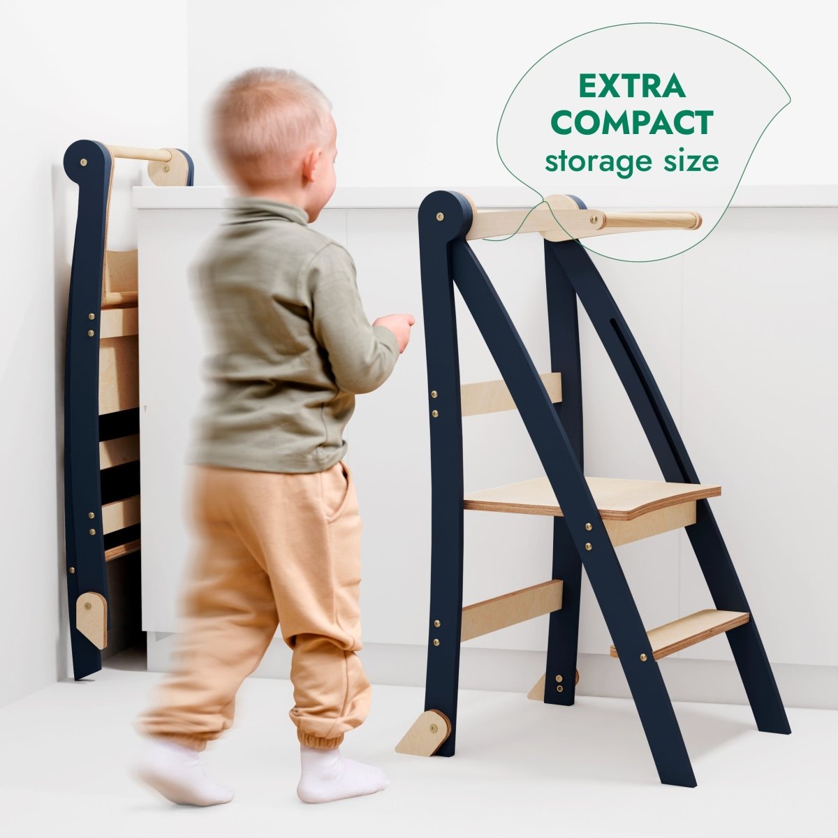 A toddler stands near a graphite and wood Toddler Tower step stool for kids, which is folded for storage against the wall. Text reads EXTRA COMPACT storage size.