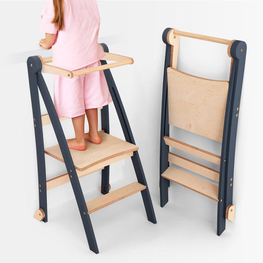 A child in pink stands at the counter on the Toddler Tower: Step Stool for Kids – Graphite. Next to her, a folded version of the same tower showcases its convenient collapsible design. Both towers combine graphite and natural wood accents, making this step stool a practical and stylish choice for little kitchen helpers.