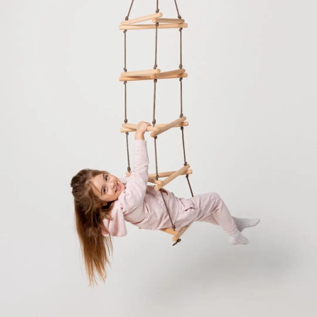 A young girl in a light pink outfit hangs and smiles while climbing on a Triangle rope ladder for kids, a developmental climbing toy, against a plain white background.