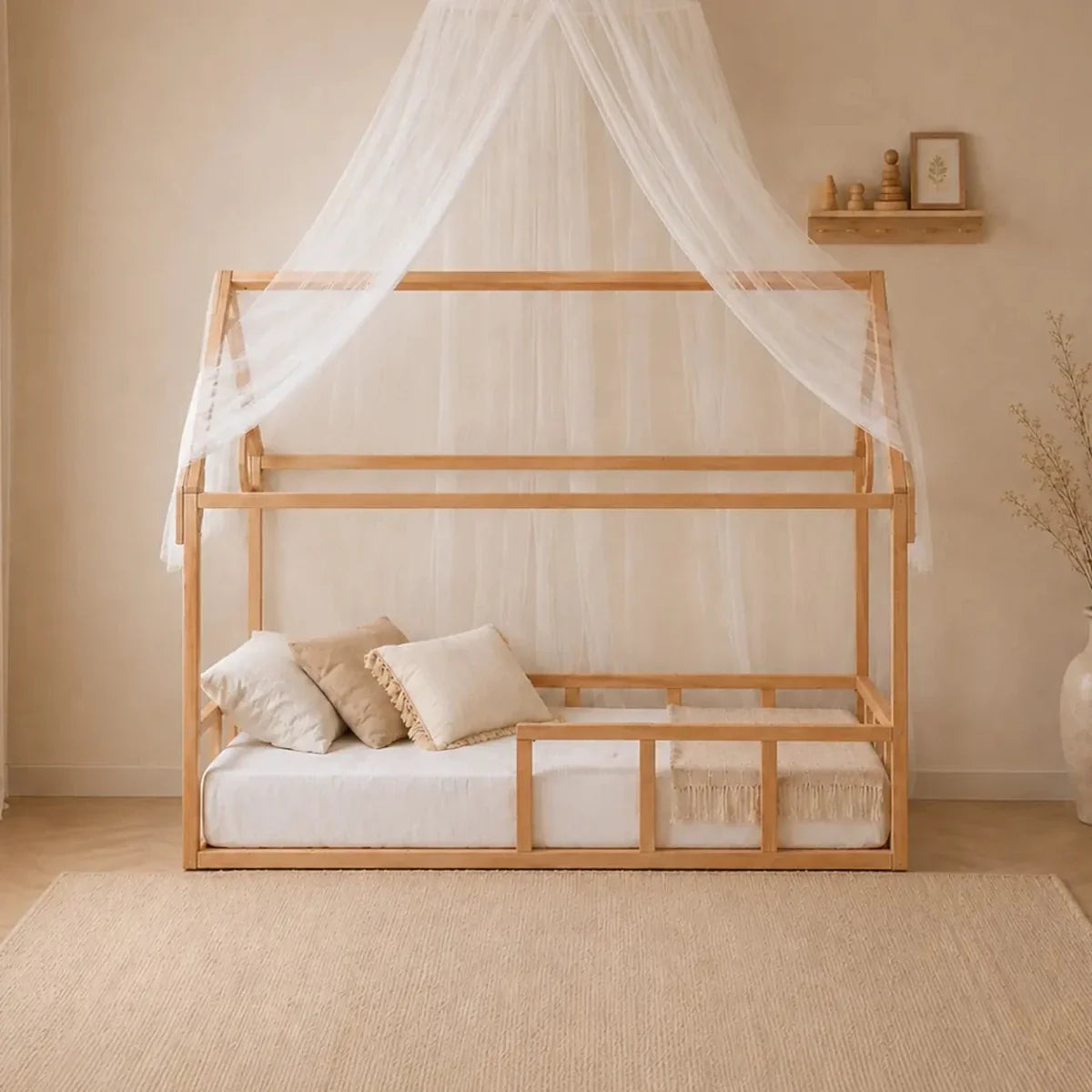 A Twin Size Montessori House Bed for Kids with Fence, crafted from solid beech wood, is styled with white bedding, neutral pillows, and a sheer canopy netting. The room complements the minimalist bed with beige walls, a small decorative shelf, and a large vase filled with dried branches.