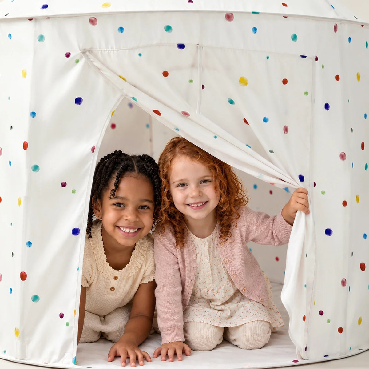 Two young girls smiling while sitting inside a white Starry Indoor Play Tent for Toddlers patterned with colorful polka dots.