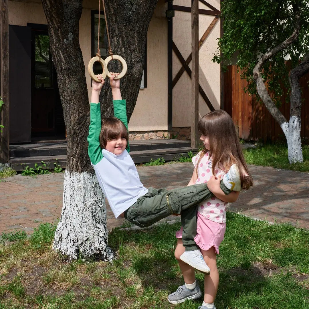 A boy smiles at the camera while hanging from Wooden gymnastic rings for kids attached to a tree, enjoying an active afternoon outdoors. Beside him, a girl stands and supports his legs, both smiling in the sunny backyard surrounded by trees and a house—the perfect setting for physical development and outdoor fun.