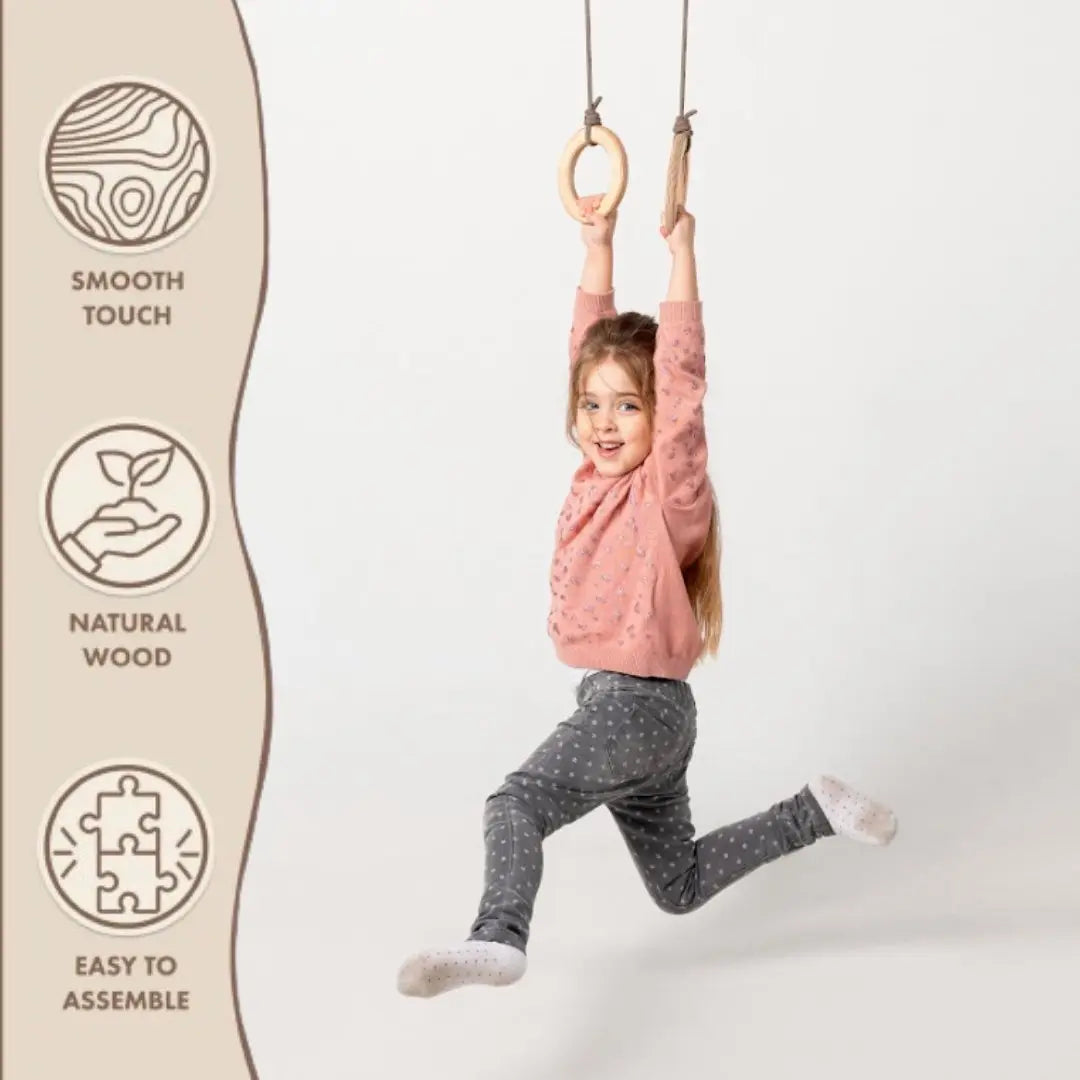 A young girl wearing a pink sweater and gray pants swings from Wooden gymnastic rings for kids. Icons on the left highlight: smooth touch, natural wood, and easy to assemble. Perfect for encouraging children’s fitness and physical development. The background is white.