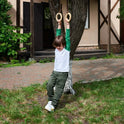 A young child in green pants and a white shirt hangs from Wooden gymnastic rings for kids attached to a tree in the yard, smiling and playing outdoors near a house—an activity that encourages physical development and boosts children’s fitness.