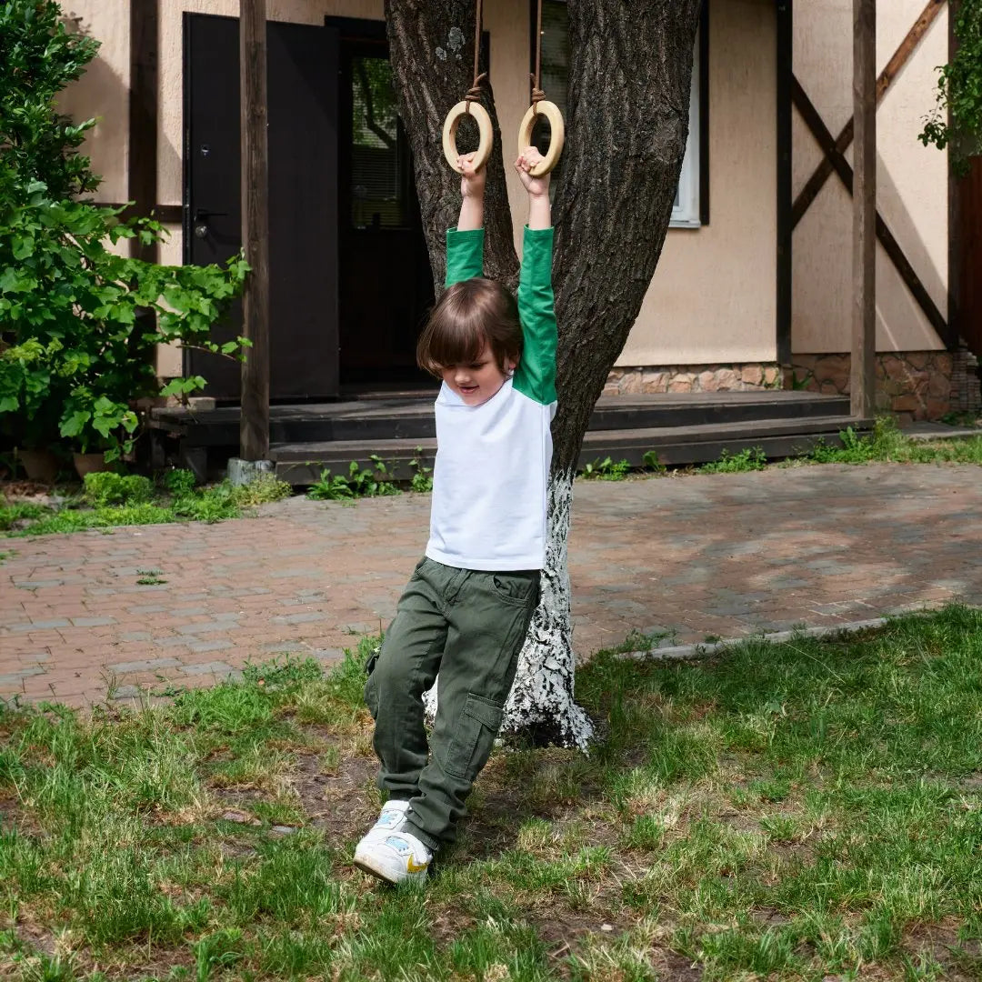 A young child in green pants and a white shirt hangs from Wooden gymnastic rings for kids attached to a tree in the yard, smiling and playing outdoors near a house—an activity that encourages physical development and boosts children’s fitness.