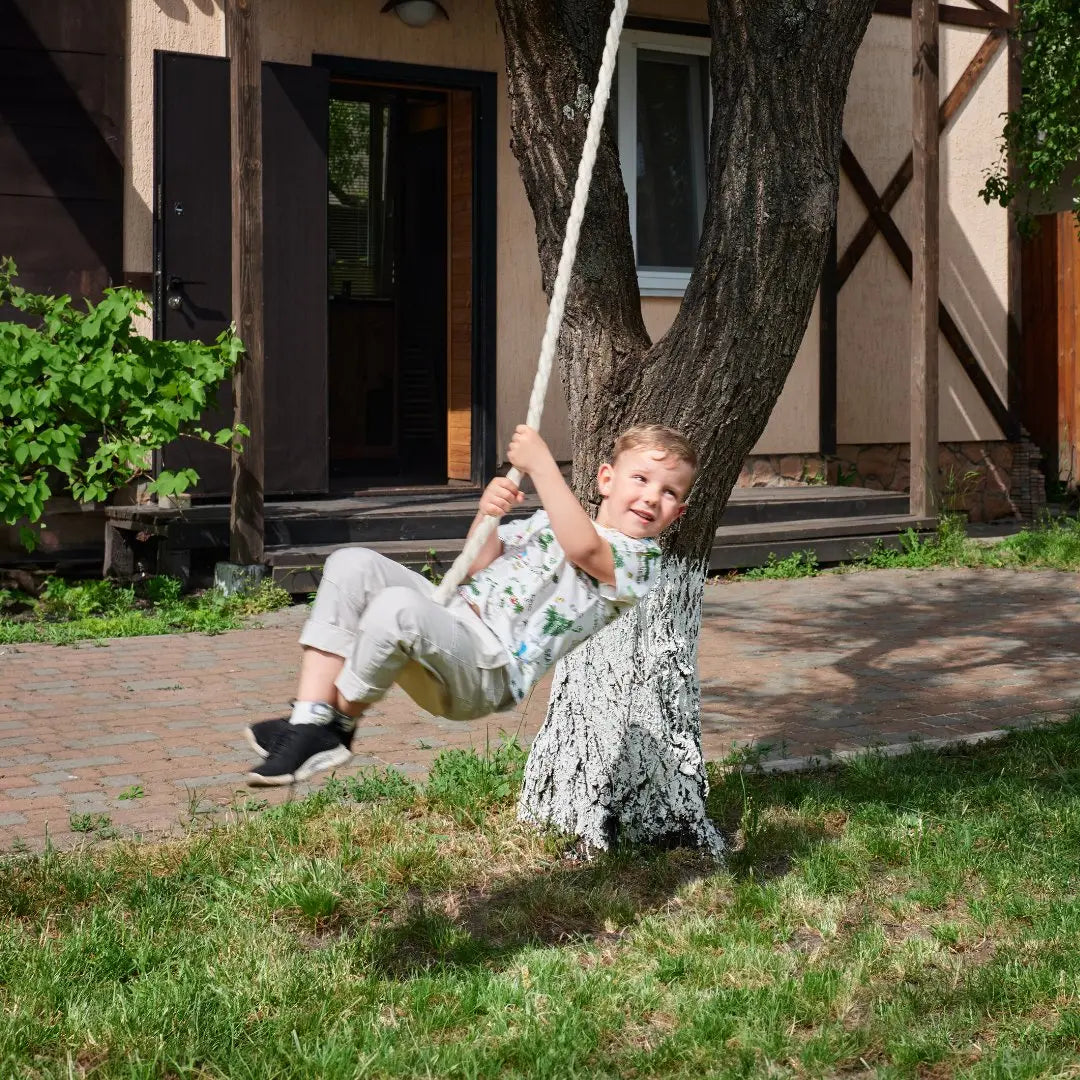 A young boy in light clothes swings on a Wooden rope swing for kids tied to a tree in a sunny yard with grass, near a house with an open door.