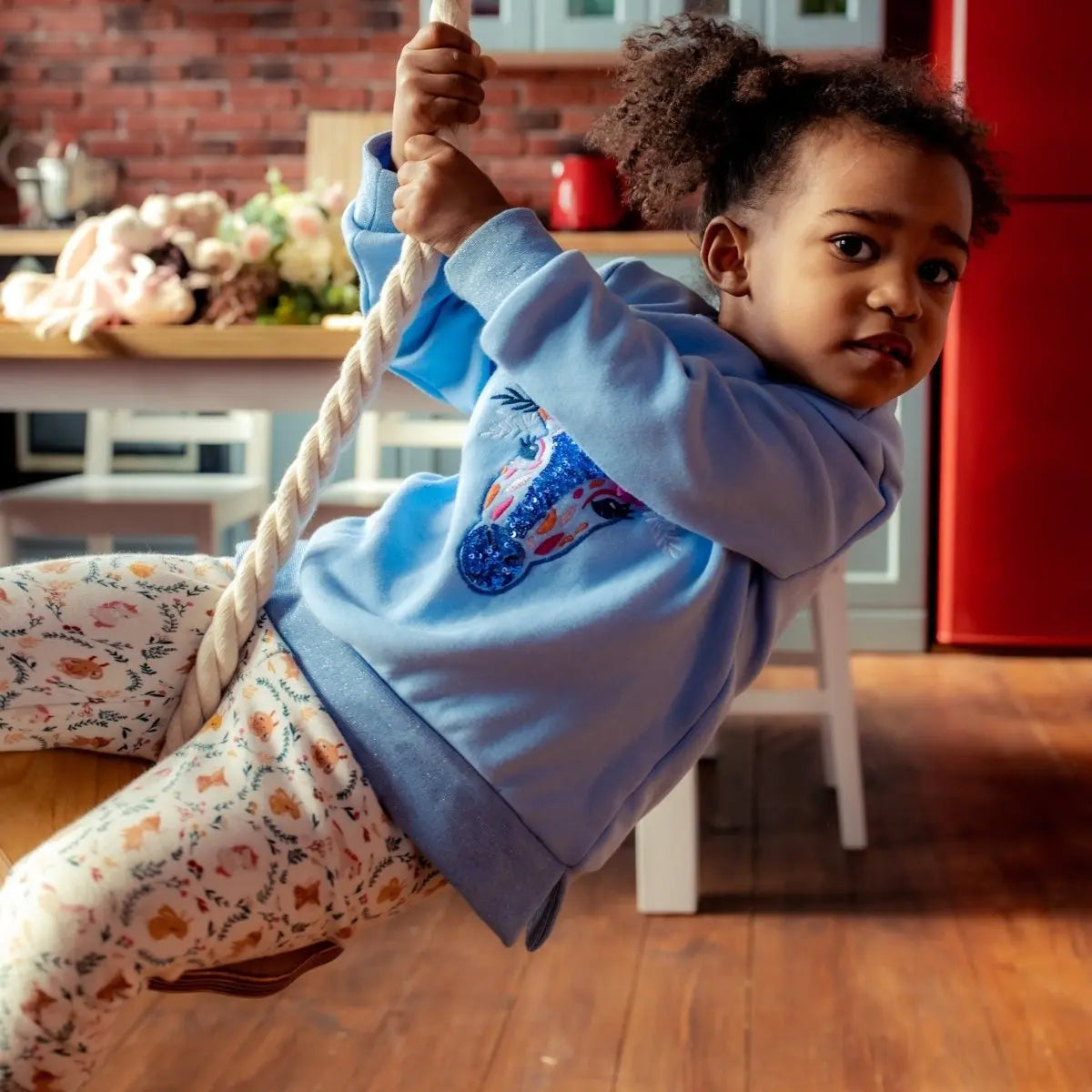 A young child with curly hair, dressed in a blue sweatshirt and floral leggings, swings on a Wooden rope swing for kids indoors, with a kitchen and dining area visible in the background, creating the atmosphere of an indoor gym.