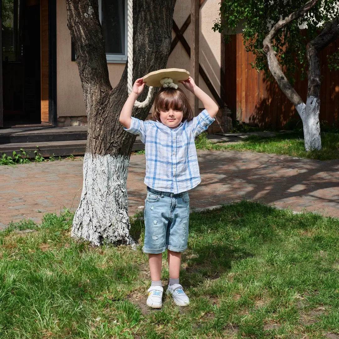 A young child in a plaid shirt and denim shorts stands on grass, holding a hat above their head in a sunny yard with trees and a house in the background—perfectly ready to enjoy the Wooden rope swing for kids.